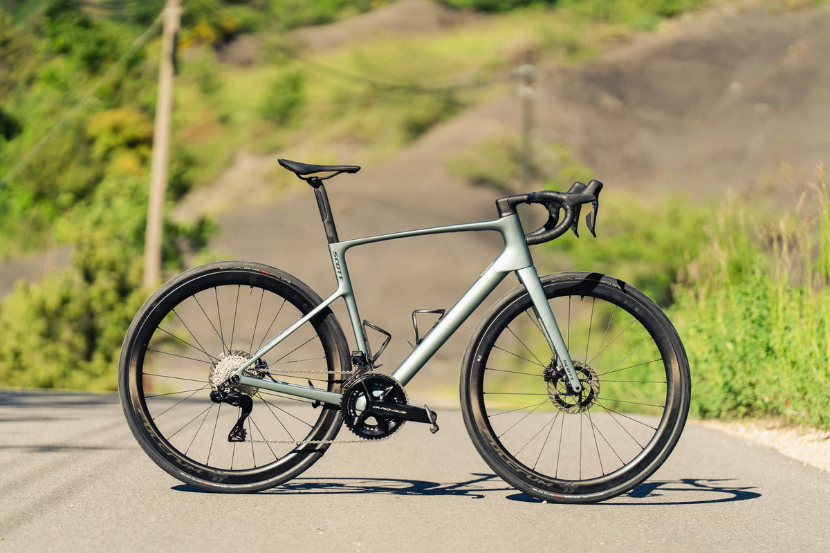 Ein silbergraues Scott Addict Rennrad steht seitlich auf einer Landstraße mit grünem Hintergrund und leichter Steigung.